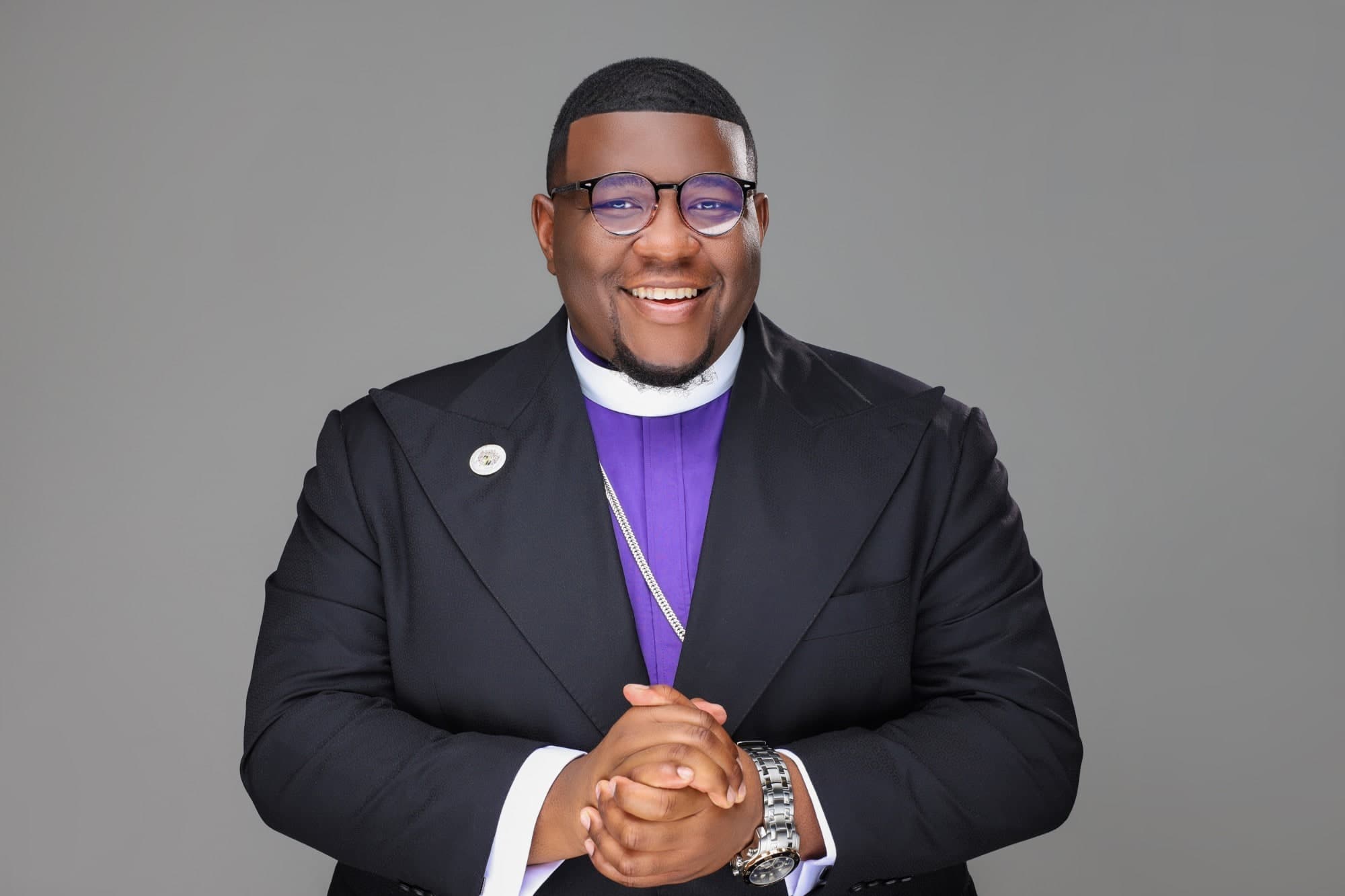 Overseer Jakeem Bowman smiling in a black suit over a purple clerical shirt and white collar, hands clasped