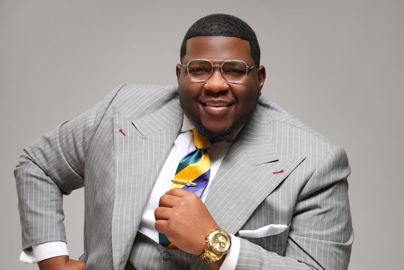Overseer Jakeem Bowman smiling in a grey pinstripe suit with a striped tie and gold watch