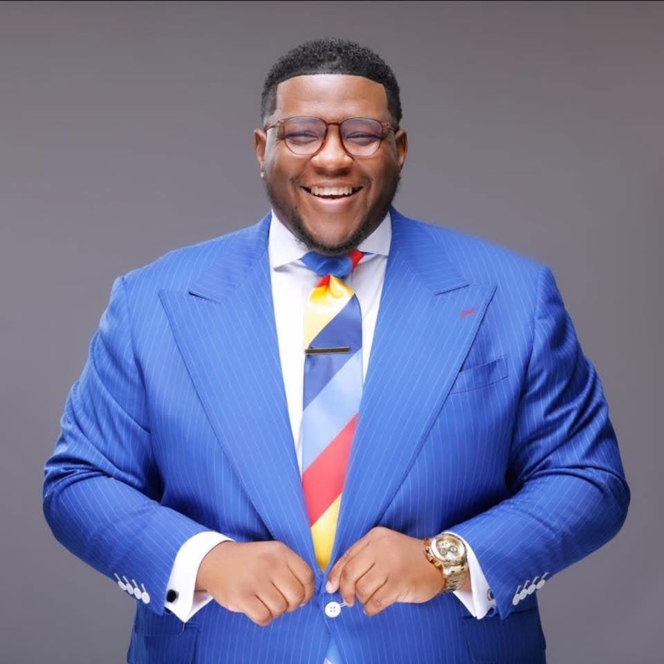 Overseer Jakeem Bowman laughing as he buttons a royal blue pinstripe suit jacket